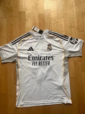 Real Madrid Mbappé matchtröja - Snygg vit Real Madrid matchtröja från Adidas med svarta detaljer och gula inslag. Tryck med Mbappé och nummer 10 på ryggen, samt klubbmärke och sponsorlogga på bröstet. Tillverkad i lätt och ventilerande material, perfekt för fotbollsträning eller match.