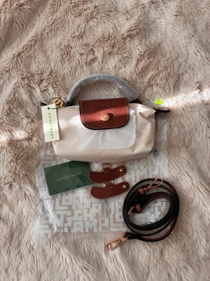 Beige handväska från Longchamp - Snygg beige handväska från Longchamp med bruna detaljer i läder och guldfärgade metalldelar. Väskan har ett klassiskt lock med tryckknapp, kort handtag och avtagbar axelrem. Perfekt för dig som gillar stilrena accessoarer med exklusiv känsla.