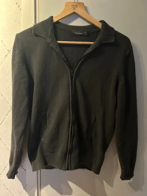 Mörkgrön tunnare cardigan från Dressmann - Mörkgrön tunnare cardigan  från Dressmann med ribbad krage, muddar och nederkant. har två fickor framtill och är tillverkad i mjukt stickat material. Perfekt för lager på lager och enkel att matcha med olika outfits.