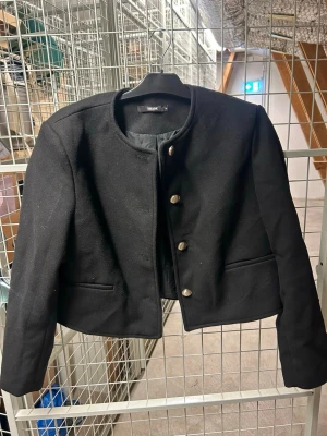 Svart kavaj med rund hals från Bik Bok - Stilren svart kavaj från Bik Bok med rund halsringning och fyra dekorativa knappar framtill. Perfekt för en trendig och clean look.