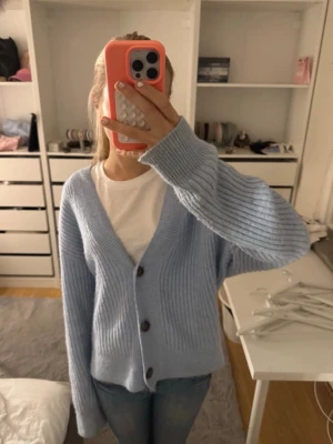 Ljusblå ribbstickad kofta - Mysig ljusblå kofta med ribbstickat mönster och stora svarta knappar framtill. Koftan har en avslappnad passform, v-ringning och långa ärmar. Perfekt att slänga över en t-shirt för en chill look.