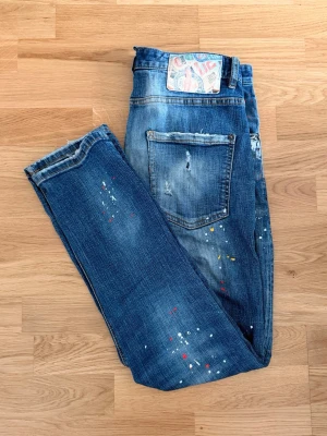 Dsquared2 jesns storlek 48 (M) - Dsquared2 med färgstänk i vitt, gult och rött. Storlek 48 (M). Obs, jeansen har en liten reva på baksidan (se bild) som ej syns vid användning, går dock att lappa ihop med denimlappar för typ 50kr som finns att köpa på ÖoB, Lindex och liknande.