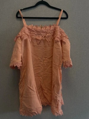 Persikofärgat nattlinne med spets - Sött nattlinne i persikofärg med tunna axelband och vackra spetsdetaljer vid halsringning, ärmslut och nederkant. Lös passform och mjukt tyg som känns skönt mot huden. Perfekt för mysiga kvällar hemma.