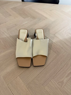 Beige slip-in sandaler från Zara - Stilrena slip-in sandaler från Zara i beige med bred rem och fyrkantig tå. Sandalerna har en chunky klack och öppen häl, vilket ger en modern look. Tillverkade i konstläder med mjuk innersula för extra komfort.