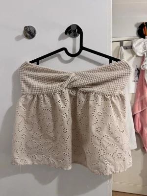 Beige axelbandslös spets tubtopp - Söt beige axelbandslös tubtopp med virkad överdel och broderade blommor i spets på hela plagget. Toppen har en twistdetalj framtill och är perfekt för varma dagar. Lätt och luftig känsla med romantiska vibbar.