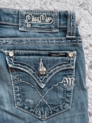 Miss Me bootcut jeans med brodyr - Säljer ett par blå bootcut jeans från Miss Me med snygga kontrastsömmar, broderade detaljer och dekorativa nitar på bakfickan. De är bootcut! Jeansen har en slitning mellan benen men det är INGET hål utan endast slitenhet💞 man ser inte vid användning. Midjemått=36cm innerbenslängd=81/82cm