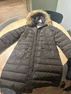 Moncler dunjacka med päls  - Snygg brun dunjacka från Moncler med huva och avtagbar päls. Jackan har två stora fickor framtill, dragkedja och tryckknappar. Lång modell som håller dig varm under kalla vinterdagar. Klassisk Moncler-logga på insidan. Storleken är S-M. Jackan är nyligen kemtvättad. 