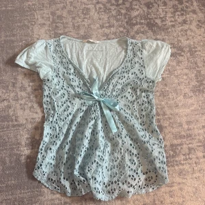 Ljusblå spetsblus med rosett - Ljusblå blus från Odd Molly med korta puffärmar, v-ringning och ett genombrutet spetslager. Framtill sitter en dekorativ rosett och materialet är mjukt och luftigt för en romantisk känsla.