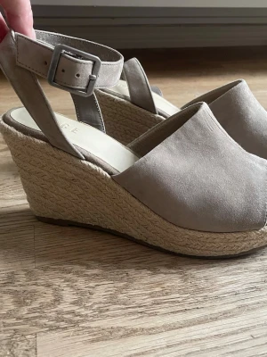 Beige kilklackar i mocka - Säljer ett par snygga beige/grå kilklackar från PACE. Skorna har öppen tå, justerbart ankelband med spänne och ovandel i mocka. Kilklacken är klädd i flätad jute för en trendig espadrille-look. Perfekta för sommarens alla outfits! Klacken är 9 cm men de är bekväma att gå i då de går upp även där fram. 