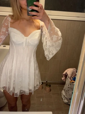 Vit studentklänning - Supersöt vit kort klänning med transparenta, vida spetsärmar och v-ringad byst med knyt. Klänningen har ett lätt utsvängt kjolparti och fina blommiga spetsdetaljer över hela tyget. Perfekt för dig som vill ha en romantisk och trendig look. Skriv för fler bilder💞