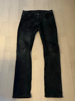 Svarta slim fit jeans - Säljer ett par svarta nudie grin tim med klassisk femficksdesign och normal midja. Jeansen har en lätt tvättad look och raka ben. Perfekta för en stilren och enkel outfit.