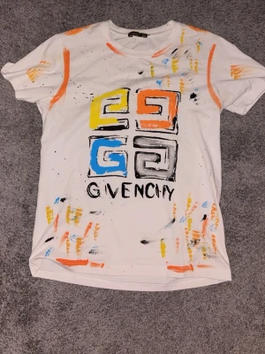 Vit Givenchy t-shirt med färgstänk - Säljer en vit t-shirt från Givenchy med färgstänk i orange, blått, gult och svart. Tryck på bröstet med grafiskt mönster och Givenchy-logga. T-shirten har rund hals och korta ärmar. Perfekt för dig som gillar streetwear och unika plagg.
