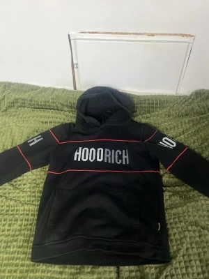 Svart hoodie från Hoodrich XL - Svart hoodie från Hoodrich i storlek XL (13-15 år). Hoodien har stor logga på bröstet, röda linjer över bröst och armar samt tryckta siffror och bokstäver på ärmarna. Utrustad med huva och stor magficka. Perfekt för en streetwear-look.