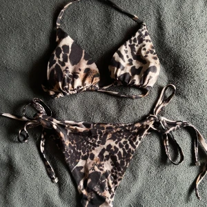 Leopardmönstrad bikini med knyt - Snygg leopardmönstrad bikini med trekantsöverdel och knyt i både topp och trosa. Bikinin har svarta och bruna toner och är perfekt för sommaren. Materialet känns mjukt och följsamt, troligtvis syntet. Perfekt för strandhäng eller poolparty! Ungefär A-B kupa och XS-S nederdel! ❤️