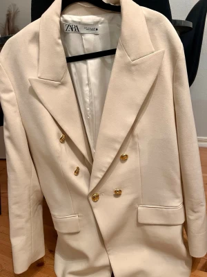 Krämvit kavaj från Zara med guldknappar - Stilren krämvit kavaj från Zara med dubbelknäppning och sex dekorativa guldknappar framtill. Klassisk oversized passform med breda slag och två fickor. Perfekt för att lyfta vilken outfit som helst med en elegant touch.