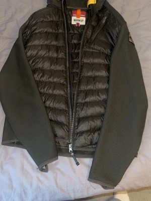 Parajumper cardigan - Snygg cardigan från Parajumpers med quiltad front och släta, mörkgrå ärmar. Jackan har dragkedja, huva och ett gult spänne vid halsen. Märkets logga sitter på vänster. Pris kan diskuteras vid snabb affär.