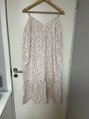 Blommig sommarklänning med smala axelband - Luftig långklänning med smala axelband och v-ringning. Klänningen är vit med små blommiga mönster i flera färger som rosa, blått och gult. Perfekt för varma dagar och har en flowy passform. Tillverkad i mjuk bomull som känns skön mot huden.