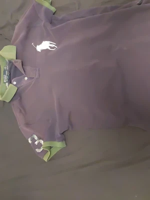 Ralph Lauren pikétröja - Säljer en mörkt lila pikétröja från Ralph Lauren med gröna detaljer på krage och ärmslut. Tröjan har en stor vit logga på bröstet och en rutmönstrad siffra 3 på ena ärmen. Klassisk pikéknäppning med två knappar och normal passform.