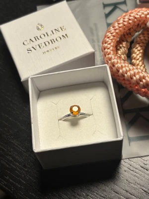 Silverring med orange sten Caroline Svedbom - Hej!🌸 Säljer en Snygg silverfärgad ring från Caroline Svedbom med en rund, orange sten i mitten och små gnistrande stenar längs bandet. Ringen har en elegant och modern design som verkligen sticker ut. Perfekt för dig som vill ha något unikt och färgglatt. Aldrig använd!      Nypris 395 kr💕