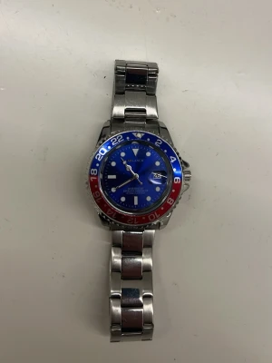 Arlanch klocka med blå och röd ring - Stilren klocka från Arlanch med blå urtavla och en tvåfärgad ring i blått och rött. Klockan har en robust länk i silverfärgad metall och tydliga markeringar. Datumvisning vid klockan tre och sportig design som sticker ut. Den är lite förkortad men tror den passar till dom flesta skriv din om du är intresserad eller nåt du undrar.