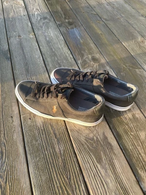 Svarta Polo Ralph Lauren sneakers - Snygga svarta sneakers från Polo Ralph Lauren med klassisk låg profil och vit sula. Skorna har canvasmaterial och den ikoniska broderade loggan i gult på sidan. Perfekta för en avslappnad och stilren look.