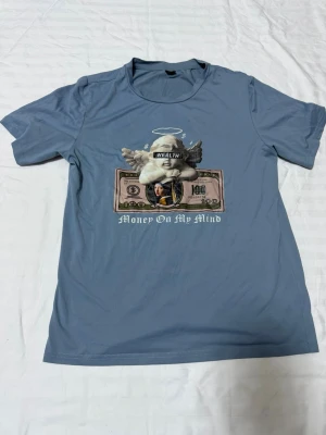 Blå t-shirt med tryck från SHEIN - Blå t-shirt från SHEIN med coolt tryck av en ängel, sedel och texten 'Money On My Mind' på bröstet. T-shirten har rund hals och korta ärmar. Materialet känns mjukt och lätt, perfekt för en avslappnad stil.