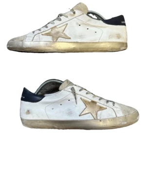 Golden Goose vita sneakers  - Golden Goose sneakers i vitt läder med beige stjärna på sidan och mörkblå häl. Skorna har grå snörning, perforerade detaljer och slitna effekter på sulan. Klassisk låg modell med retrovibe och cool streetstil.