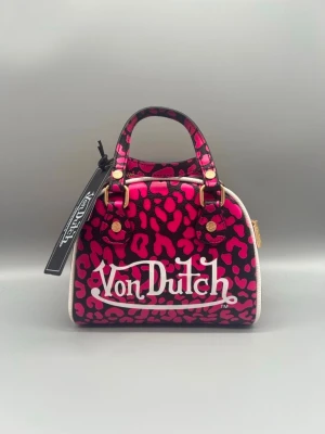 Von Dutch rosa leopard handväska - Cool handväska från Von Dutch med rosa och svarta leopardmönster, vit logga framtill och vita kanter. Väskan har guldfärgade metalldetaljer, dubbla handtag och dragkedja upptill. Perfekt statement piece för dig som gillar att sticka ut.