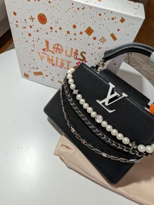 Iouis Vuitton Capucines - Design: En del av den ikoniska Capucines-linjen, namngiven efter Iouis Vuittons första butik i Paris (öppnad 1854).Den är djupt svart med en tydlig textur i lädret, vilket ger ett sofistikerat och robust intryck. Väskan utmärker sig genom sin eleganta dekorationskedja som hänger i en båge över framsidan; kedjan består av tre rader med stora vita pärlor, en klassisk metallkedja och en modernare länk med fyrkantiga element.