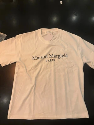  Maison Margiela t-shirt - Vit t-shirt från Maison Margiela med klassisk rund hals och svart tryck med logotypen 'Maison Margiela PARIS' på bröstet. Enkel och stilren design som passar perfekt till jeans eller shorts. Tillverkad i mjuk bomull för en skön känsla.