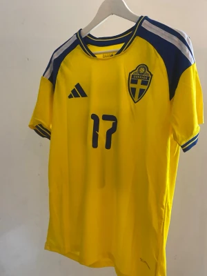 Sverige fotbollströja Adidas gul och blå #17 gyökers  - Gul fotbollströja från Adidas med Sveriges landslagsmärke, nummer 17 och namnet Gyökeres på ryggen. Tröjan har blå detaljer på axlar och ärmslut samt Climalite-material för bra ventilation. Perfekt för att bära på matchdag och för ett stundande vm💛💙
