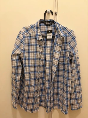 Blårutig skjorta från Åhléns - Snygg blårutig skjorta från Åhléns i bomull. Skjortan har klassisk krage, långa ärmar och knäppning framtill. Perfekt för en avslappnad och stilren look. Färgerna går i blått, vitt och beige.