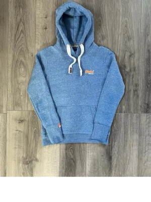 Blå hoodie från Superdry - Snygg blå hoodie från Superdry med vit dragsko i huvan och klassisk Superdry-logga broderad i orange på bröstet. Hoodien har en stor magficka och mjukt material som känns skönt mot huden. Perfekt för en avslappnad och cool stil.