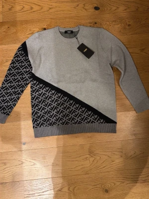 Grå stickad Fendi-tröja med mönster - Snygg grå stickad tröja från Fendi med svart diagonal logomönstrad detalj och en helärm i svart/grått mönster. Rund halsringning och ribbade muddar. Perfekt för dig som gillar exklusiva och trendiga plagg.
