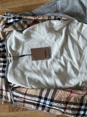 Grå Burberry hoodie - Snygg grå hoodie från Burberry med dragkedja och den ikoniska beigea, svarta och röda rutan på insidan. Mjuk bomullskänsla och huva. Perfekt för dig som vill ha en stilren och lyxig look med en touch av klassiskt brittiskt mode.