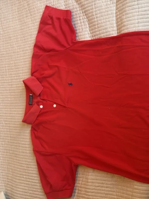 Röd pikétröja från Polo Ralph Lauren - Klassisk röd pikétröja från Polo Ralph Lauren med blå broderad logga på bröstet. Tröjan har korta ärmar, krage och två vita knappar vid halsen. Perfekt för en stilren och avslappnad look.