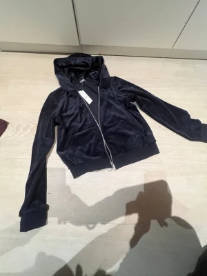  Mörkblå velourhoodie från Lindex - Mjuk och glansig svart hoodie i velour från Lindex med dragkedja framtill och huva. Perfekt för en chill och stilren look. Ribbstickade muddar vid ärmslut och nederkant. Snygg detalj med diskret broderi på ena sidan. Helt nytt skick aldrig använt den
