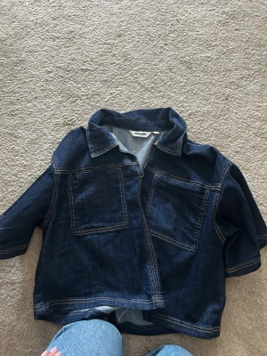 Mörkblå jeansjacka från Kappahl - Snygg mörkblå jeansjacka från Kappahl med raka linjer och två stora bröstfickor. Jackan har klassisk krage och kontrastsömmar som ger en cool look. Perfekt att slänga över en hoodie eller t-shirt.
