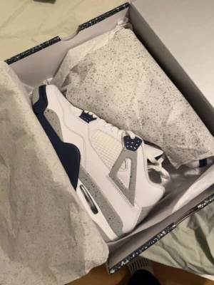 Air Jordan 4 White Navy Grey(äkta) - Snygga Air Jordan 4 sneakers i vitt läder med detaljer i marinblått och grått. Klassisk siluett med meshpaneler på sidorna, synlig Air-enhet i sulan och ikoniska plastdetaljer. Perfekta för dig som vill sticka ut med stilrena och sportiga sneakers.