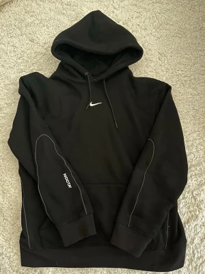 Nike NOCTA hoodie - Säljer en svart hoodie från Nike NOCTA med broderad vit Swoosh och NOCTA-logga på bröstet. Hoodien har huva med dragsko, magficka och vita detaljer längs ärmarna. Perfekt för dig som gillar streetwear och vill ha en stilren look.