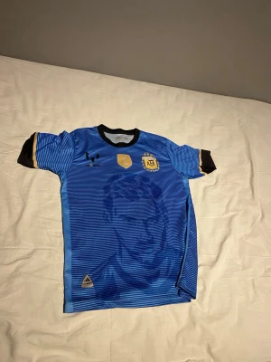 Argentina Messi matchtröja blå - Säljer en blå Argentina matchtröja från Adidas med Messi-tryck och nummer 10 på ryggen. Tröjan har svarta och gula detaljer, rund hals och porträtt av Messi på framsidan. Perfekt för dig som älskar fotboll och vill stötta ditt favoritlag.