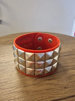 Punk armband - Ett rött punkarmband med tre rader av silverfärgade pyramidformade nitar. Armbandet stängs med tryckknappar. PU läder. Storlek 22 cm långt. Bredd 4,6 cm.