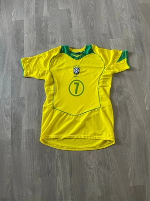 Gul Brasilien fotbollströja nr 7 - Snygg gul fotbollströja från Brasilien med gröna detaljer och nummer 7 på framsidan. Tröjan har CBF-emblemet broderat på bröstet och texten BRAZIL under. Tillverkad i lätt och ventilerande material, perfekt för träning eller match.