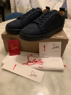 Mörkblå sneakers från Louboutin - Snygga mörkblå sneakers från Christian Louboutin i mocka med klassisk röd sula. Skorna har rund tå, svarta snören och diskret branding på sidan. Perfekta för dig som vill ha en lyxig och stilren look med en ikonisk touch.