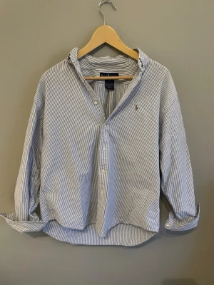 Randig skjorta från Polo Ralph Lauren - Klassisk blå- och vitrandig skjorta från Polo Ralph Lauren med broderad logga på bröstet. Skjortan har knäppning framtill, lång ärm och avslappnad passform. Tillverkad i mjuk bomull som känns skön mot huden. Perfekt för en clean och stilren look.