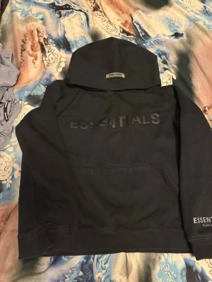 Svart Essentials hoodie Fear of God - Svart hoodie från Fear of God Essentials med stor huva och tryckt logga på bröstet samt ärmen. Klassisk känguruficka framtill och ribbade muddar. Perfekt för en streetwear-look och riktigt skön passform.