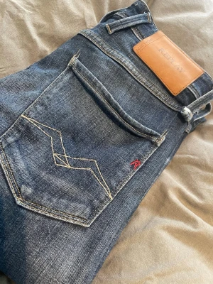 Blå Replay jeans med smal passform - Snygga blå jeans från Replay med smal passform och klassisk femficksdesign. Jeansen har slitningar och ljusa tvättade partier, samt Replay-logga på läderpatch bak. Dekorativ söm på bakfickan och liten röd detalj. Perfekta för en trendig vardagslook. 