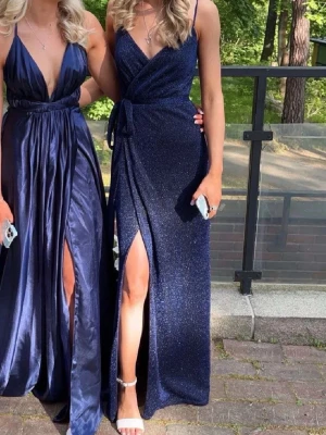 Marinblå glittrig långklänning med slits - Snygg marinblå långklänning med glittrigt tyg och omlott-design. Klänningen har smala axelband, djup v-ringning och en hög slits framtill. Knytband i midjan ger en fin passform (kan tas bort). Perfekt för dig som vill sticka ut på student eller bal. PRIS KAN DISKUTERAS 