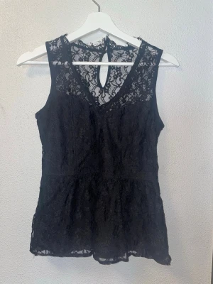 Svart spetslinne med v-ringning - Snygg svart ärmlös topp i spets med v-ringning och transparenta detaljer upptill. Toppen har en liten knapp i nacken och är perfekt för att lyfta vilken outfit som helst. Passar dig som gillar en feminin och trendig stil.