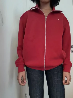 Röd zip-hoodie från Puma - Röd hoodie från Puma med dragkedja hela vägen, ribbade muddar och diskret Puma-logga på bröstet. Klassisk modell utan huva, perfekt till jeans eller joggers. Mjuk bomullskänsla och två fickor framtill.